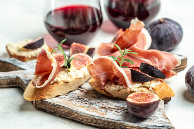 Bruschette gourmet con fichi e prosciutto su un tagliere di legno e calici di vino rosso