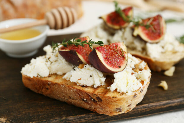 Primo piano di un tagliere di legno con bruschetta di fichi, formaggio di capra e ciotola di miele su sfondo bianco