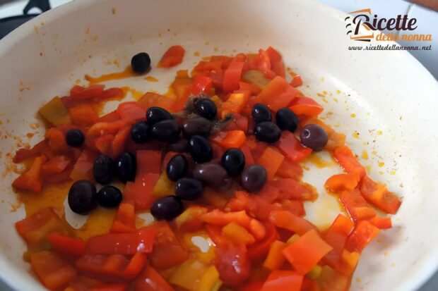 Chopped red and yellow bell peppers sautéing with black olives and anchovy fillets in a white frying pan, showing the early stage of red pepper pasta sauce preparation.