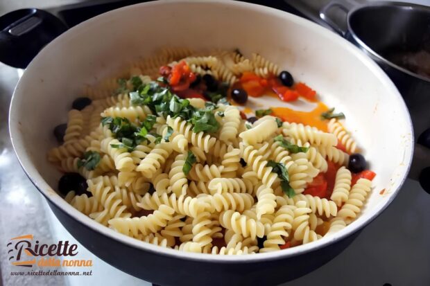 Cooked fusilli pasta tossed with sautéed red bell peppers, black olives, and fresh basil in a white pan, showing the final mixing stage of red pepper pasta sauce preparation.