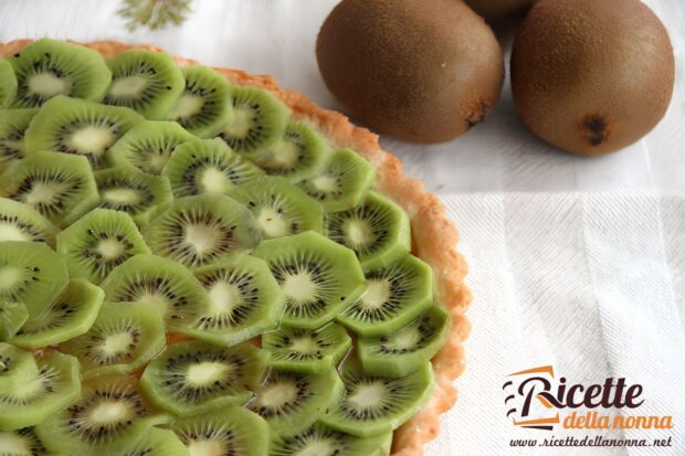 Primo piano di una crostata di kiwi con la frolla senza crema, e sopra fettine di kiwi sovrapposte. Tre kiwi interi su sfondo di tovaglia bianca
