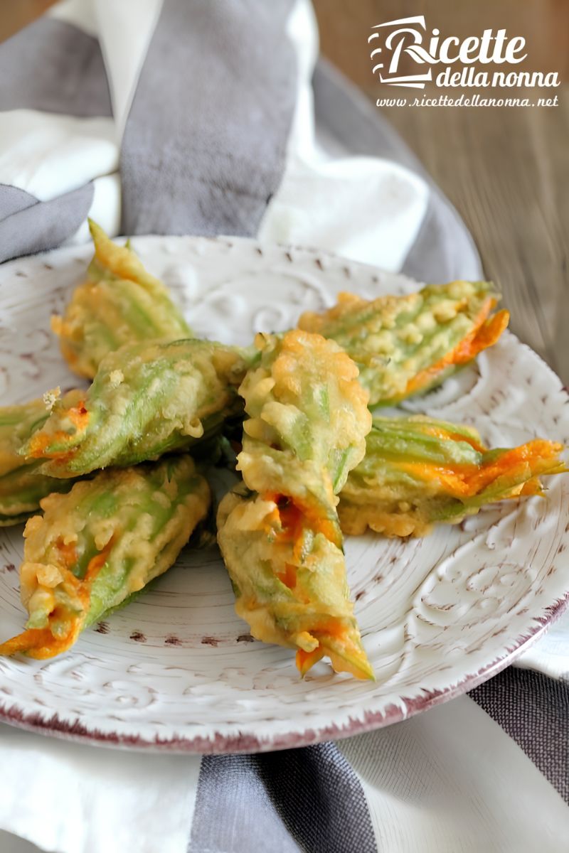 Fiori di zucca fritti in pastella croccante, disposti su un piatto decorato bianco con bordo marrone, su tovagliolo a righe bianche e grigie. Primo piano