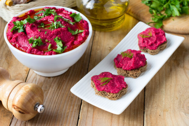 Ciotola bianca piena di hummus di barbabietola color fucsia brillante, guarnito con prezzemolo fresco e olio d’oliva, accanto a tre crostini rotondi di pane scuro spalmati con l'hummus e decorati con rametti di aneto, su un tavolo di legno.