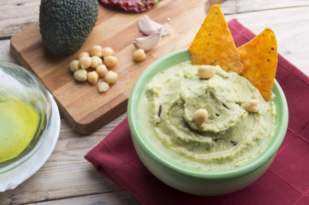 Ciotola di hummus di ceci e avocado con ceci interi e nachos, servita con olio extravergine su tagliere in legno.