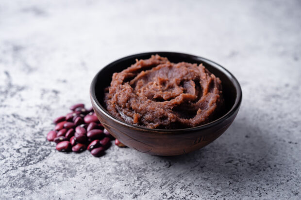 Ciotola in legno scuro riempita con hummus di fagioli rossi dalla consistenza compatta, con alcuni fagioli rossi secchi sparsi accanto su un piano chiaro in marmo.