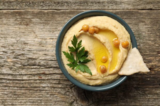 Ciotola di hummus senza tahina guarnita con ceci interi, prezzemolo e un filo d’olio d’oliva, con un pezzo di pita sul bordo, su sfondo di legno rustico.