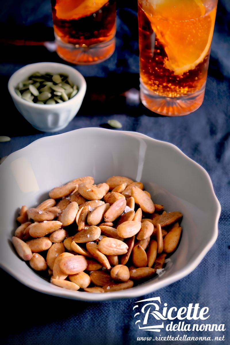 Ciotola di mandorle tostate e salate su tovaglia scura; sullo sfondo un bicchiere con uno spritz e una ciotolina di semi di zucca.