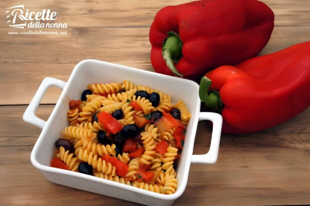 Fusilli pasta with diced red bell peppers, black olives, and light tomato dressing in a white square dish, with two fresh red bell peppers beside it on a wooden table.