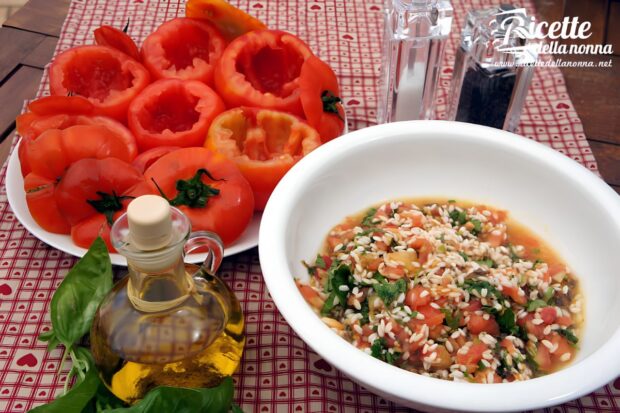 Piatto di pomodori scavati e ciotola con l'interno di riso ed altri ingredienti, su un tavolo di legno con vicino bottiglie di olio, vino e aceto