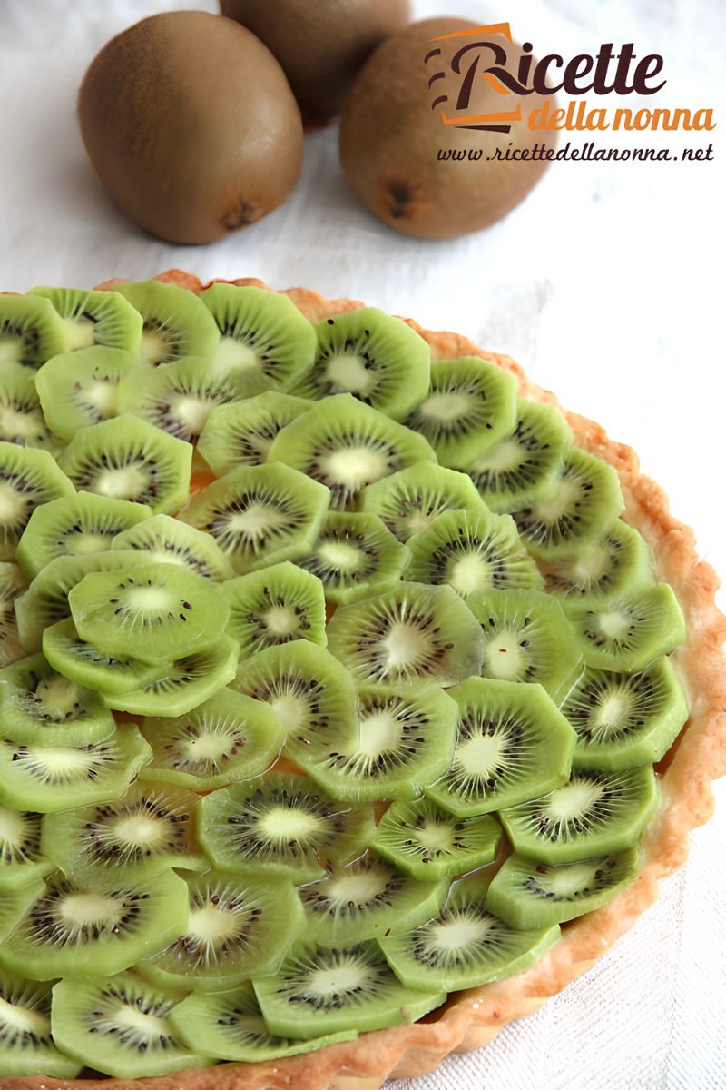 Primo piano di una crostata di kiwi con la frolla senza crema, e sopra fettine di kiwi sovrapposte. Tre kiwi interi su sfondo di tovaglia bianca