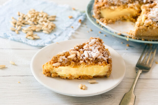 Fetta di torta alle mele e frutta secca, con crema gialla e crumble, coperta di zucchero a velo e pinoli, su un piattino bianco con una forchetta accanto. Sfondo con sparsi e la torta intera