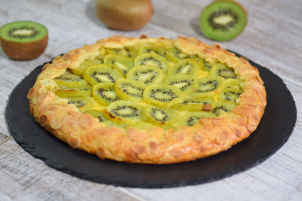 Torta di pere con crema al timo, decorata con fette di kiwi, bordo cosparso di mandorle a lamelle, servita su un piatto nerp