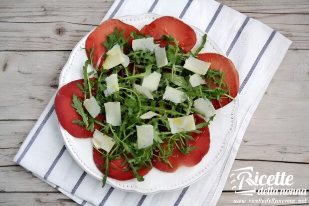 Bresaola con rucola e grana su un piatto bianco sopra una tovaglia bianca con strisce azzurre, su sfondo di legno.