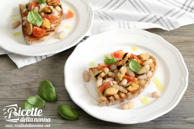 Bruschette servite su piatto bianco, condita con cozze, pomodori a pezzi, fagioli cannellini e foglie di basilico fresco, con un filo d’olio extravergine d’oliva