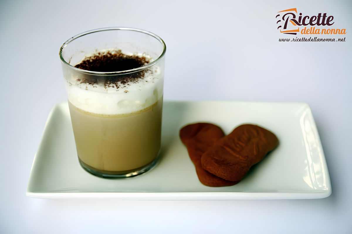 Bicchiere di panna cotta al cappuccino con panna montata e cacao in polvere, servita su un piatto bianco con due biscotti.