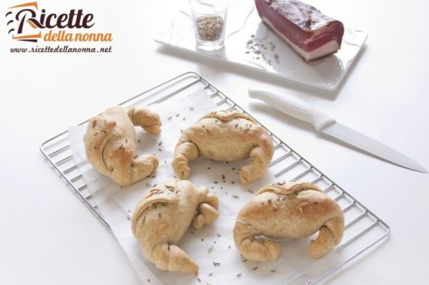 Quattro cornetti salati crescenza e speck su una gratella. Vicino un coltello e sopra lo speck su di un vassoio. Sfondo bianco