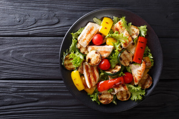 Insalata di pollo con verdure grigliate servita in un piatto nero: petto di pollo grigliato a strisce, zucchine, peperoni rossi e gialli, pomodorini ed altro