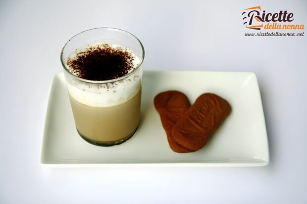 Bicchiere di panna cotta al cappuccino con panna montata e cacao in polvere, servita su un piatto bianco con due biscotti.
