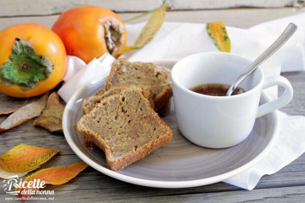 Due fette di Plumcake ai cachi servite in un piattino grigio con accanto un caffè in tazza. Due cachi sullo sfondo