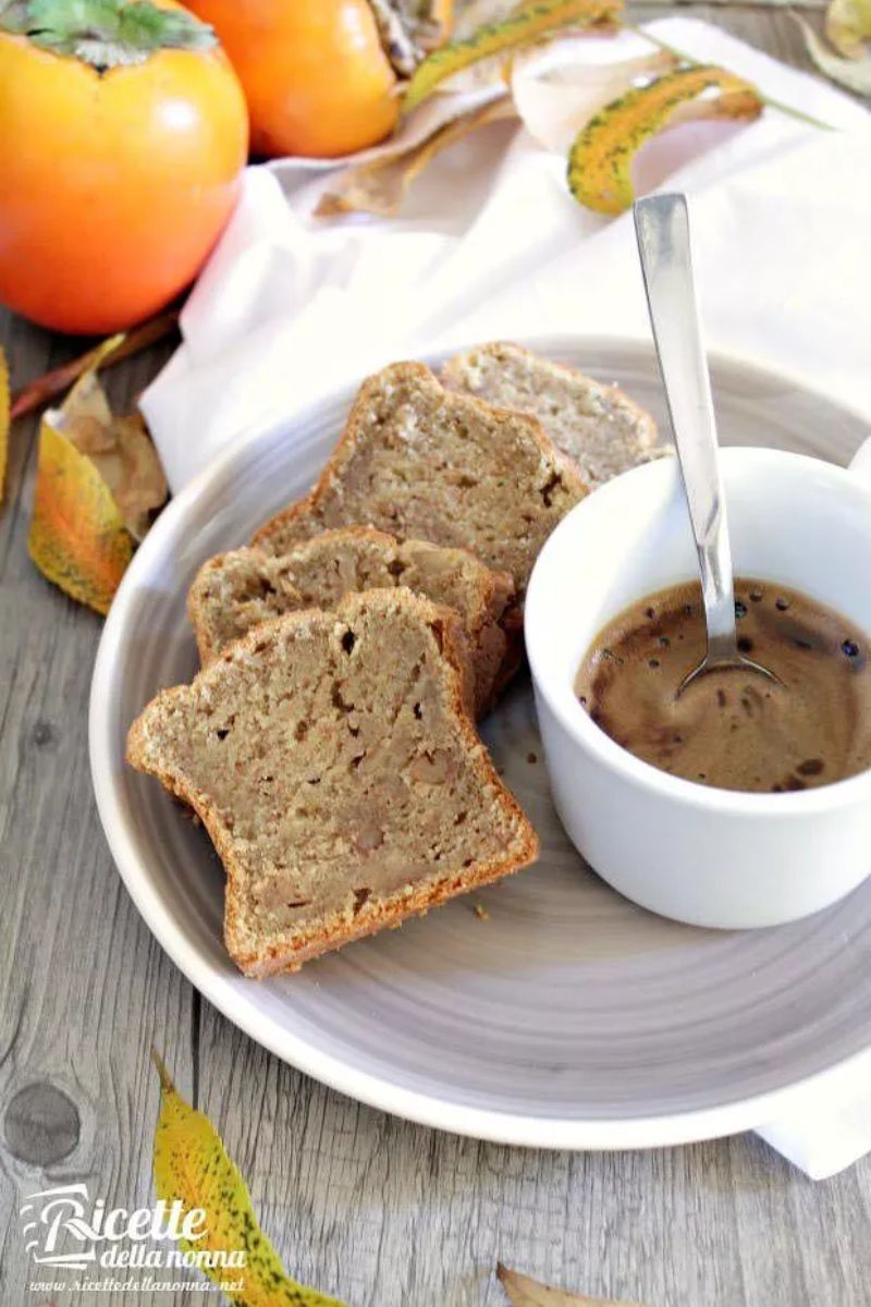 Primo piano di due fette di Plumcake ai cachi servite in un piattino grigio con accanto un caffè in tazza. Due cachi sullo sfondo