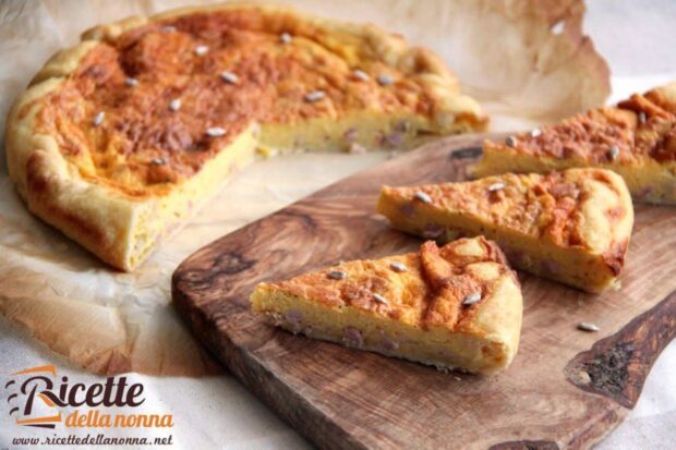 Tre fette di Quiche Lorraine su tagliere di legno e restante Quiche Lorraine sullo sfondo. Visibili pezzi di pancetta