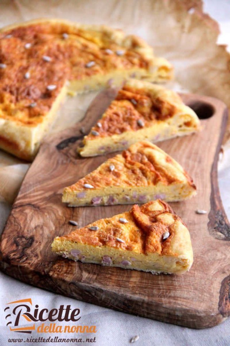 Primo piano di tre fette di Quiche Lorraine su tagliere di legno e restante Quiche Lorraine sullo sfondo. Visibili pezzi di pancetta