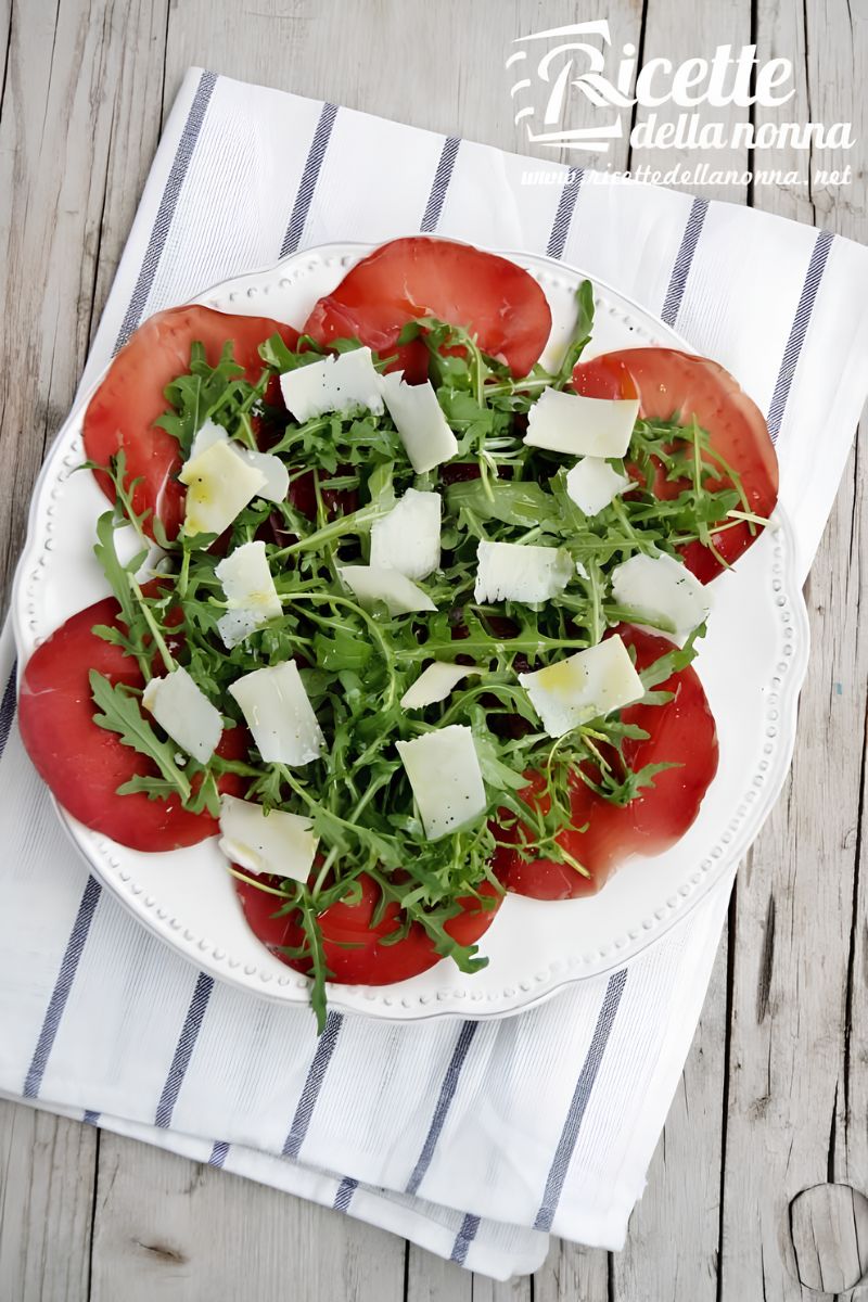 Bresaola con rucola e grana su un piatto bianco sopra una tovaglia bianca con strisce azzurre, su sfondo di legno. Primo piano