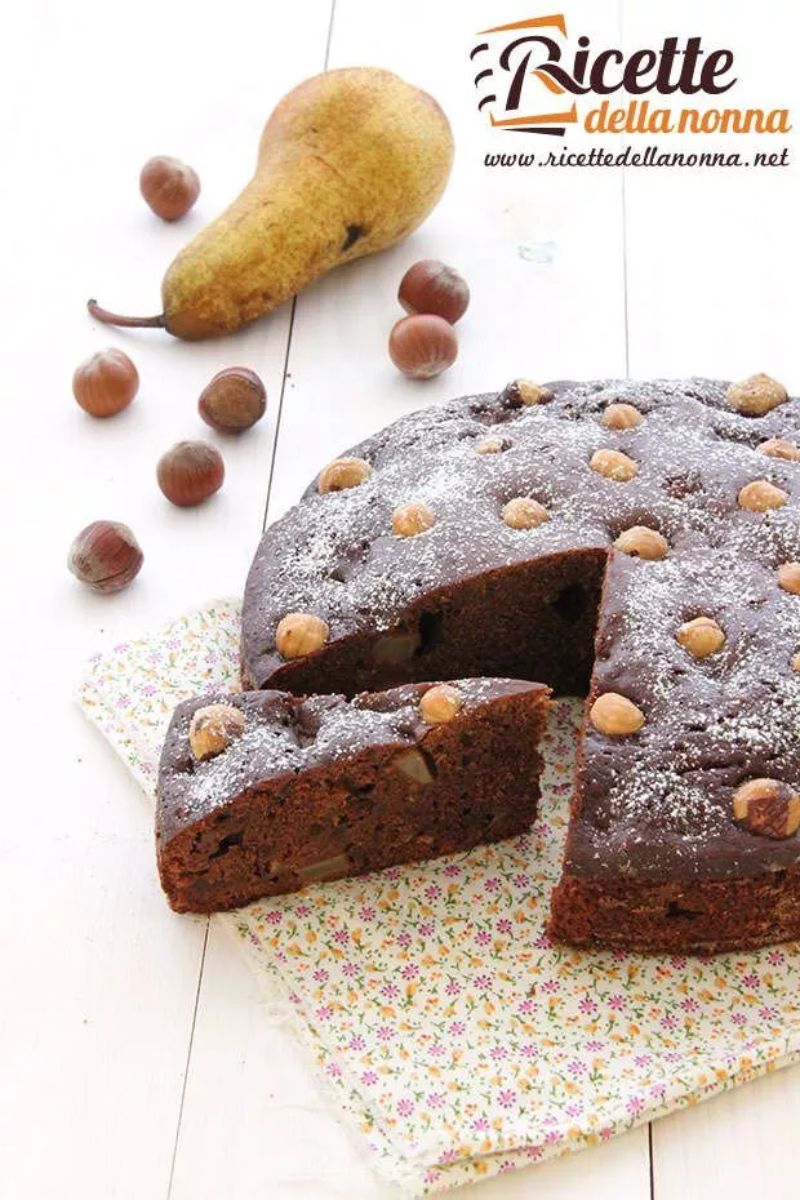 Torta cioccolato nocciole e pere, poggiata su una tovaglia fiorata. Una pera e nocciole accanto per decorazione. Sfondo di legno. Primo piano
