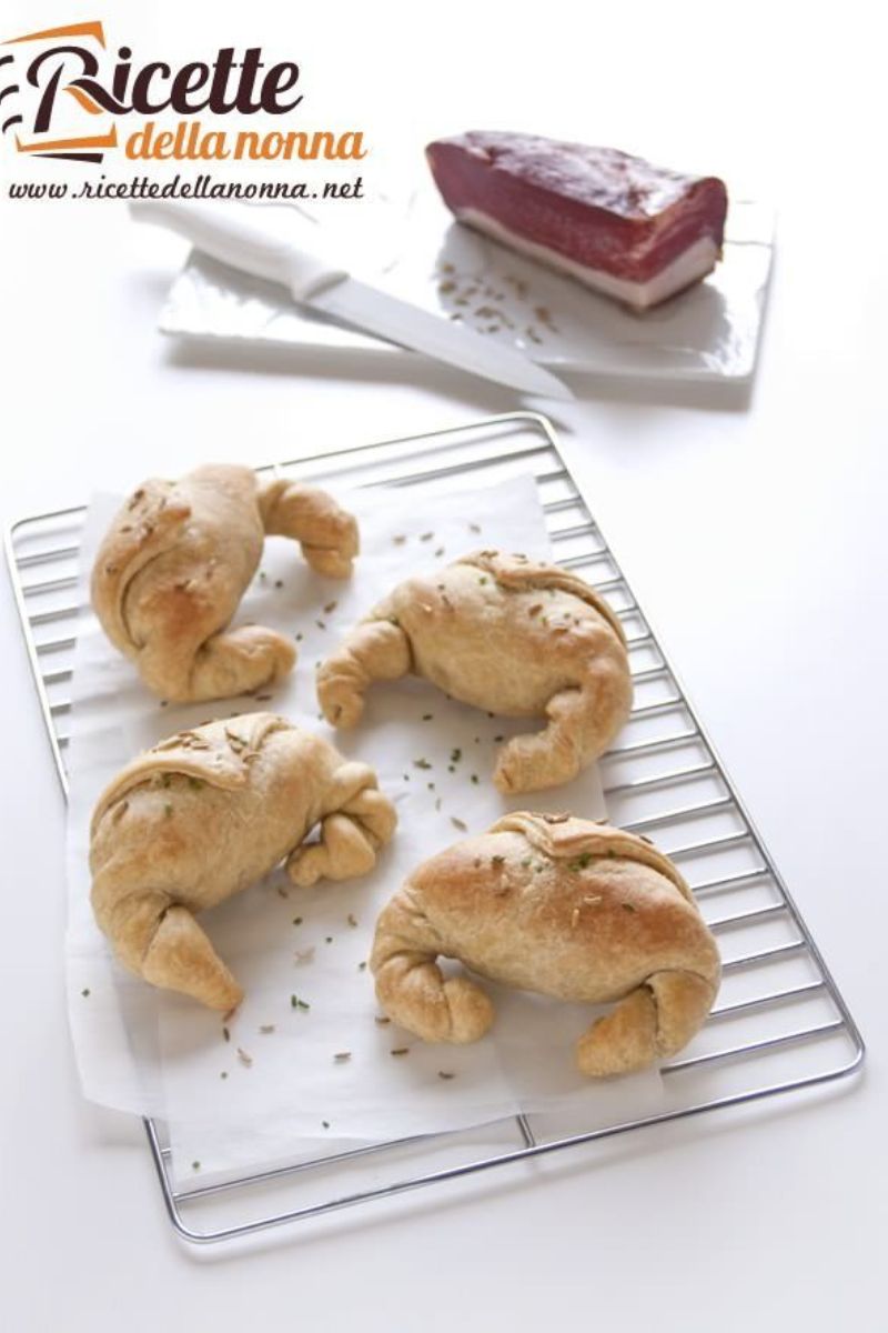 Quattro cornetti salati crescenza e speck su una gratella. Vicino un coltello e sopra lo speck su di un vassoio. Sfondo bianco