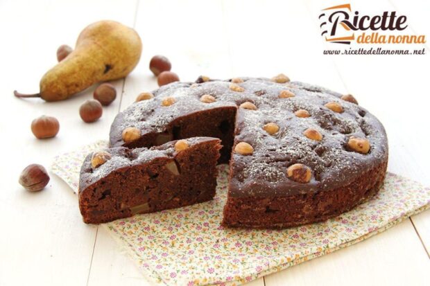 Torta cioccolato nocciole e pere, poggiata su una tovaglia fiorata. Una pera e nocciole accanto per decorazione. Sfondo di legno