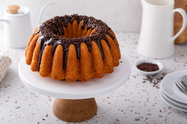 Torta pere mele e cioccolato a ciambella, decorata con glassa di cioccolato fondente e codette, messa su un’alzata bianca con base di legno