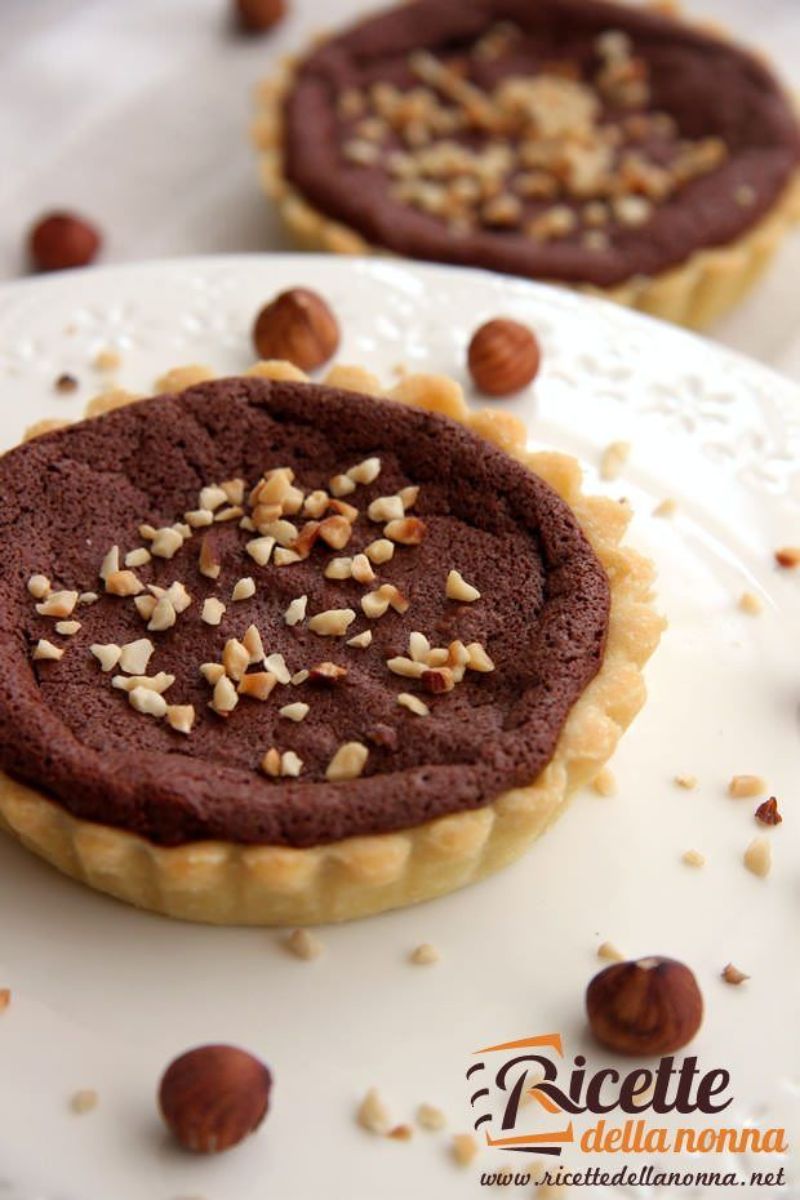2 tortine di frolla al cioccolato con base di pasta frolla, ripieno fondente e granella di nocciole, una servita su piatto bianco e decorate da nocciole intere. L'altra di sfondo