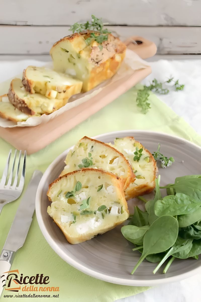 Fette di tortino di zucchine al forno con pezzi di formaggio, con vicino spinaci a decorazione, in un piatto; tortino di zucchine intero sullo sfondo. Primo piano