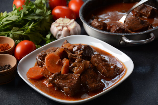 Brasato al Chianti su un piatto bianco, con accanto la pentola. Aglio e pomodori lateralmente